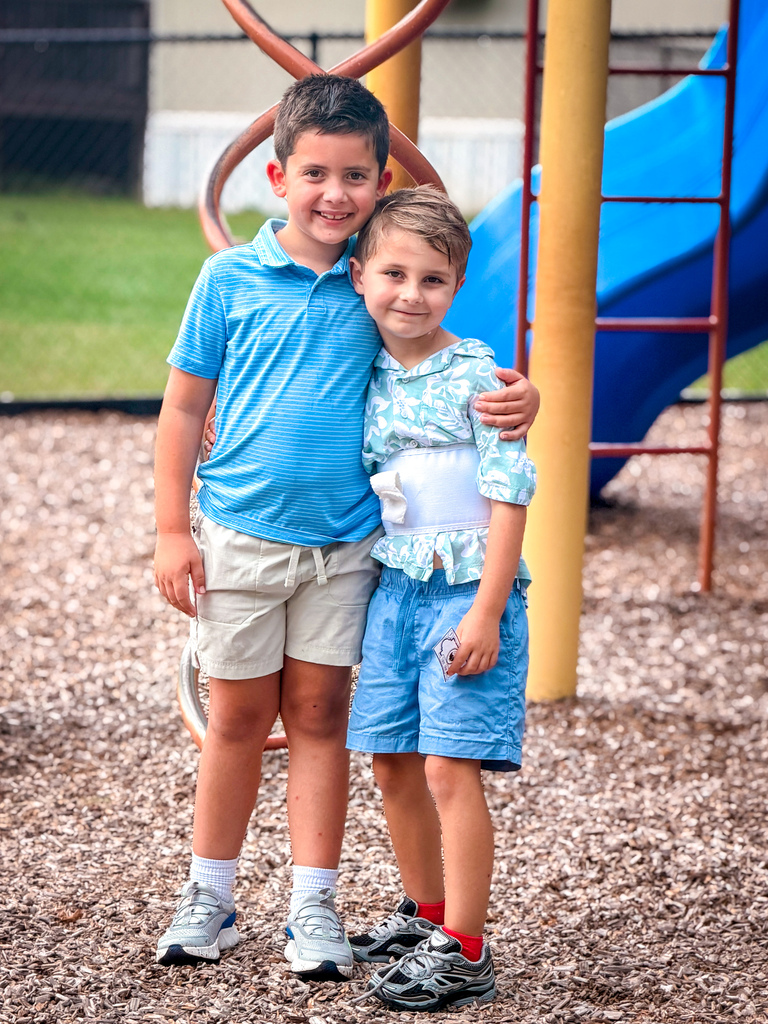Two boys, one with a brace, stand in a playground with a blue slide and yellow ladder behind them.