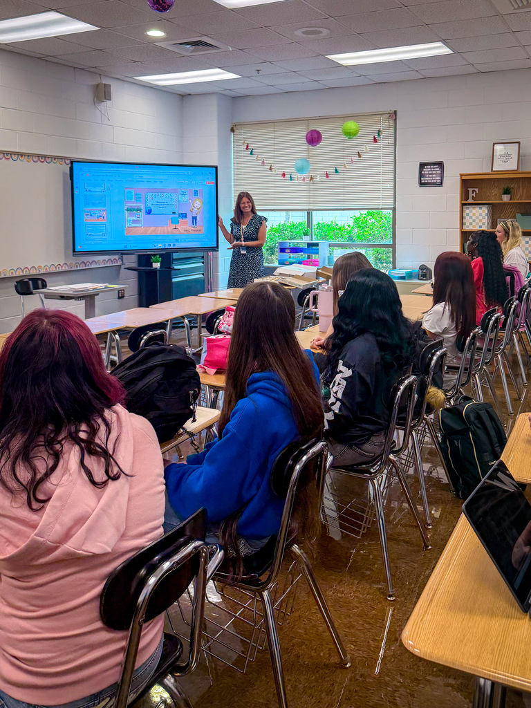 A teacher stands before a large screen, leading a class of seated students in a classroom.