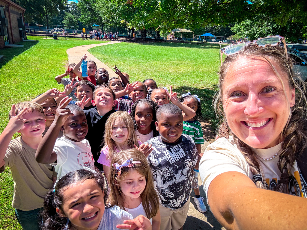 A group of children stands outside, with a smiling adult woman in the front. They pose for a picture with various gestures.