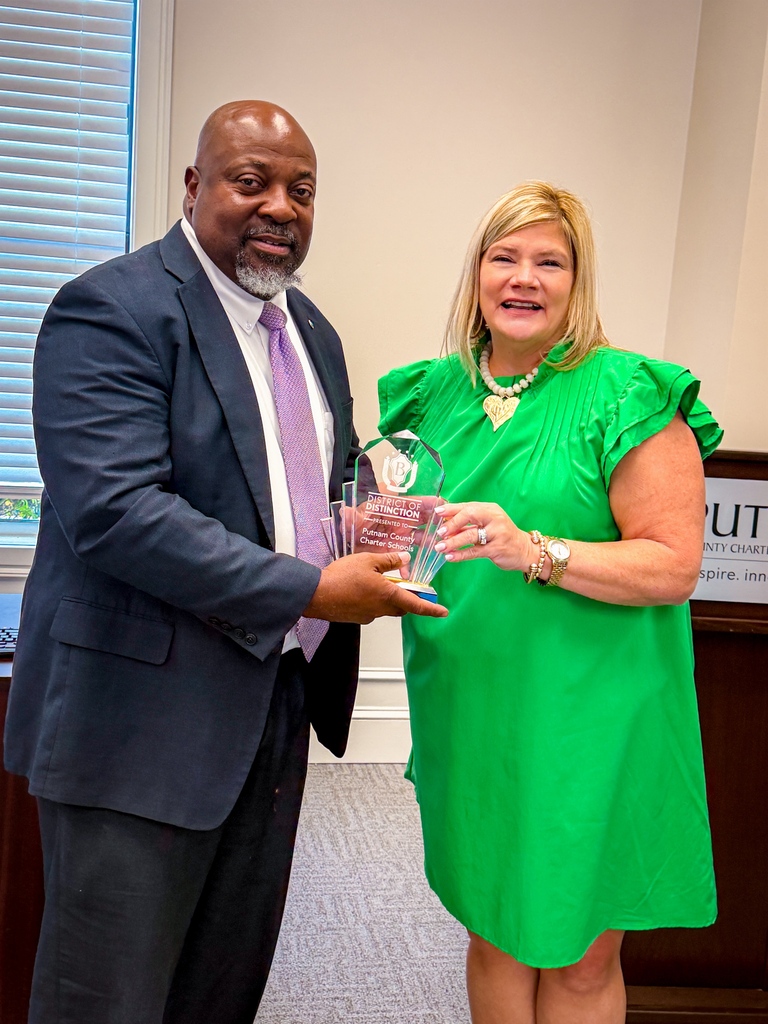 Man in a suit and woman in a green dress holding a trophy while standing in an office.