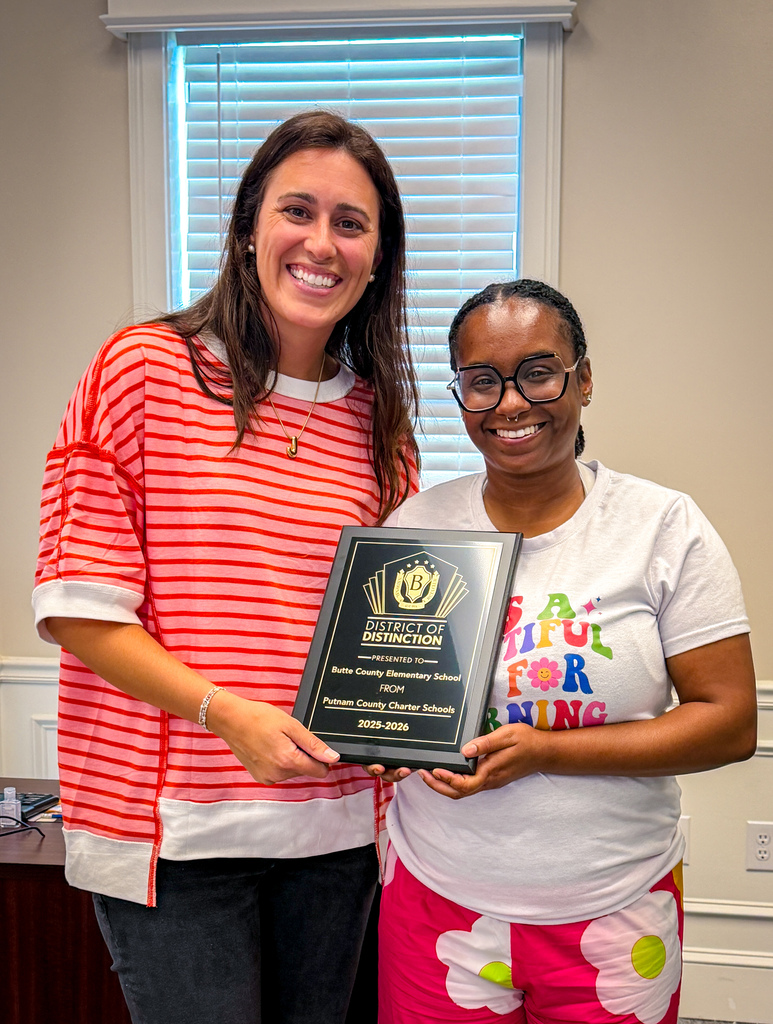 Two women holding a plaque with text. One in a red and white shirt, the other in white.