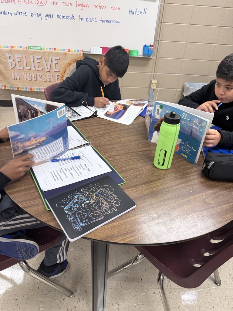 Three students at a table with books and a water bottle. A whiteboard with the word 'believe' is on the wall.
