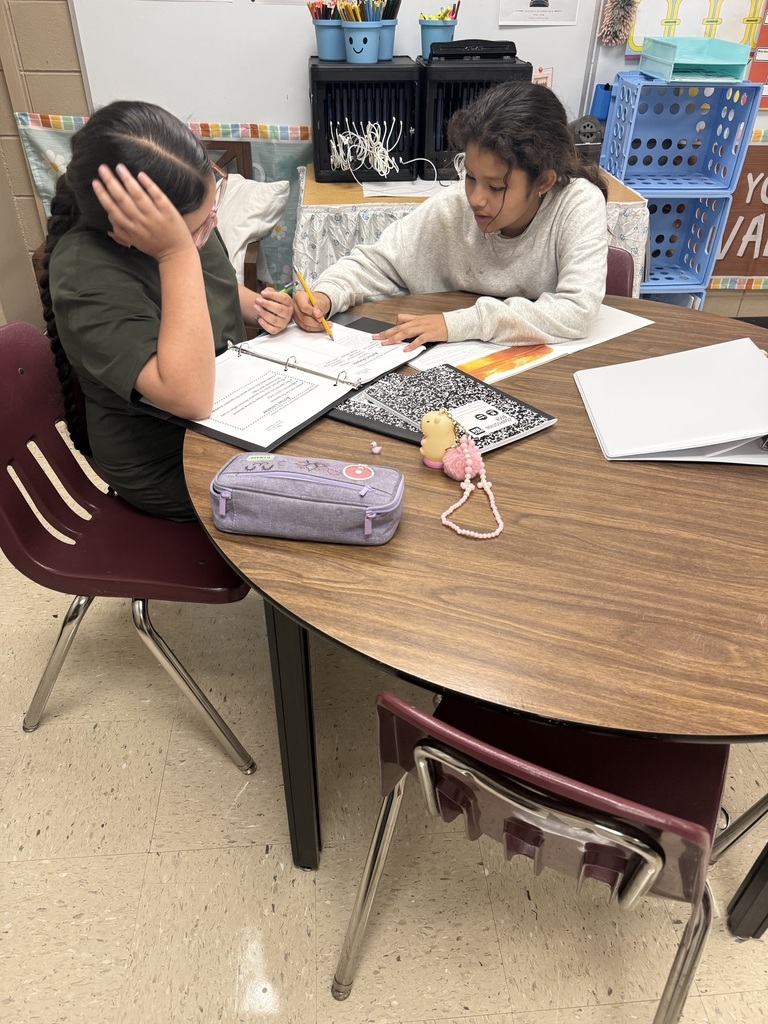 Two students seated at a table, working on papers and books, one looking concerned, other studying.