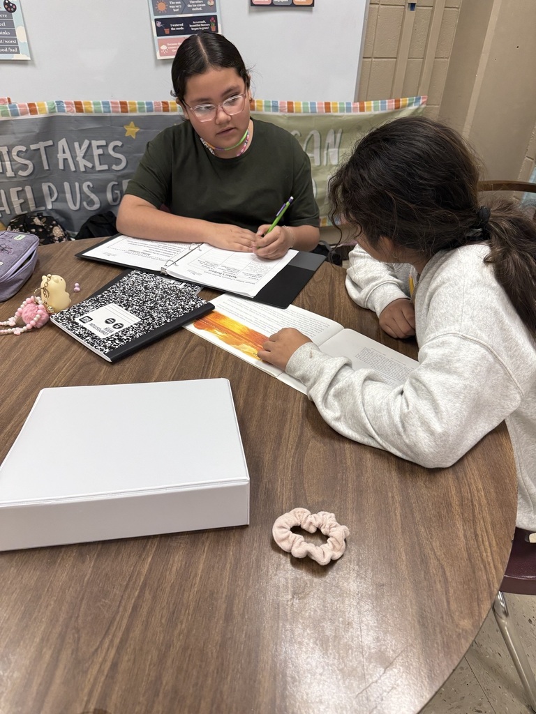 Two people sit at a table, one holding a pencil and writing while the other reads. A banner behind them reads "Mistakes help us grow."