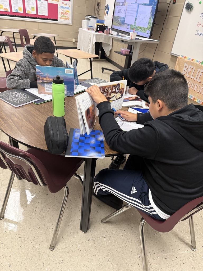 Four students in a classroom around a table with various books and a water bottle.