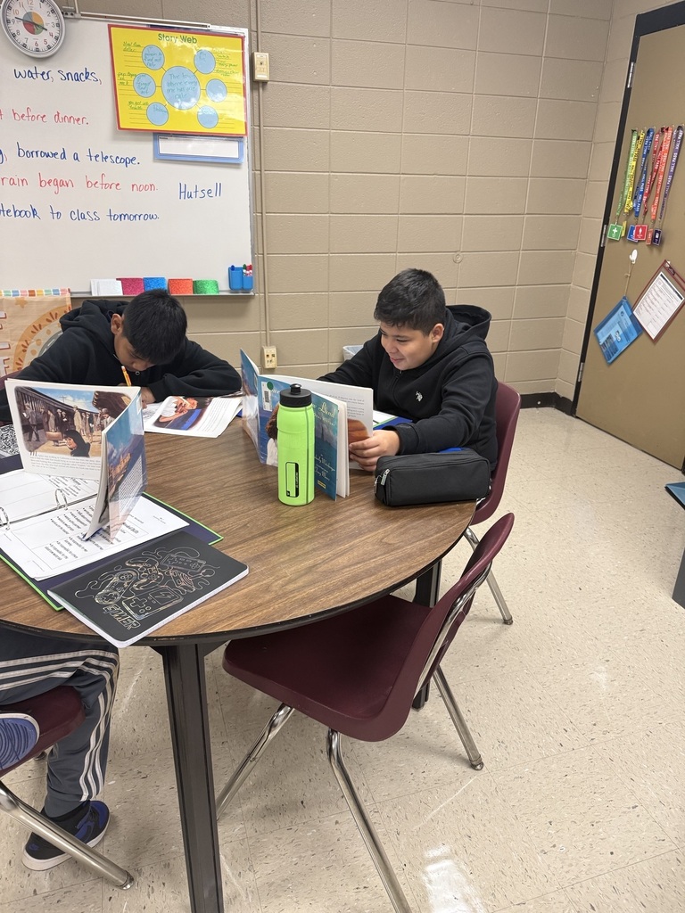 Two students sit at a table, reading from books. The table has a water bottle, papers, and a folder.
