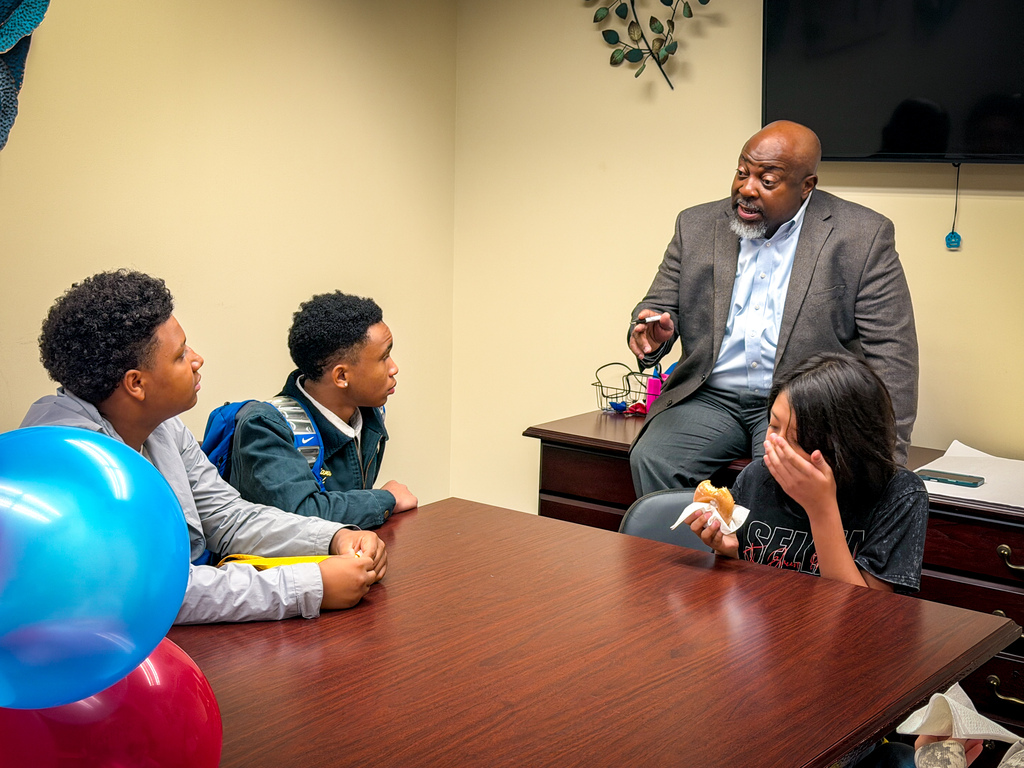Three children and an adult sit at a table with balloons. A man stands behind them. A TV and a wall decoration are visible.