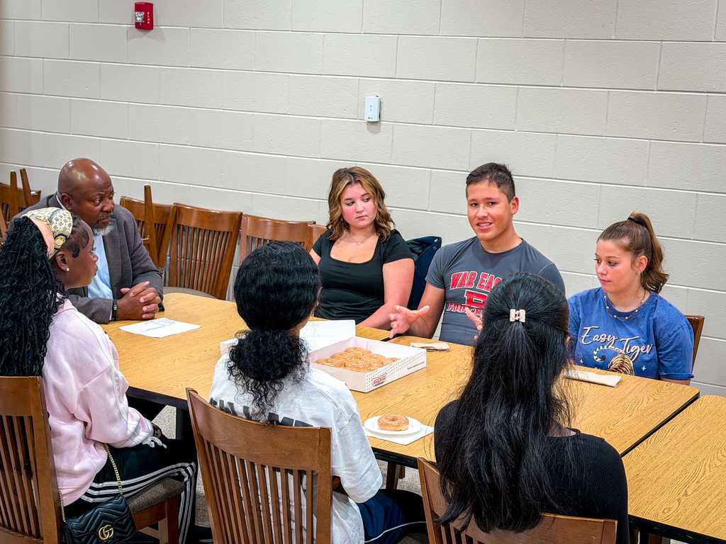 A group of people sit at a table with food in a classroom. A man speaks, while others listen.