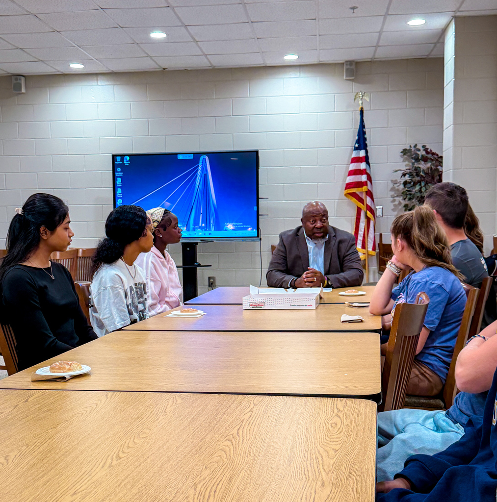 A group of people sit around tables in a room. A man sits at a table in front of a monitor.