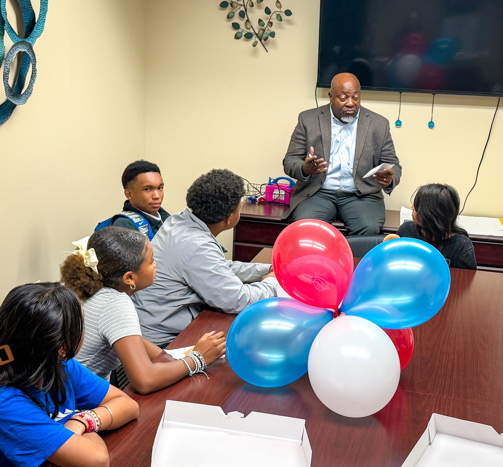 Group of people sit around a table with balloons. A man sits on a chair in front of the table.