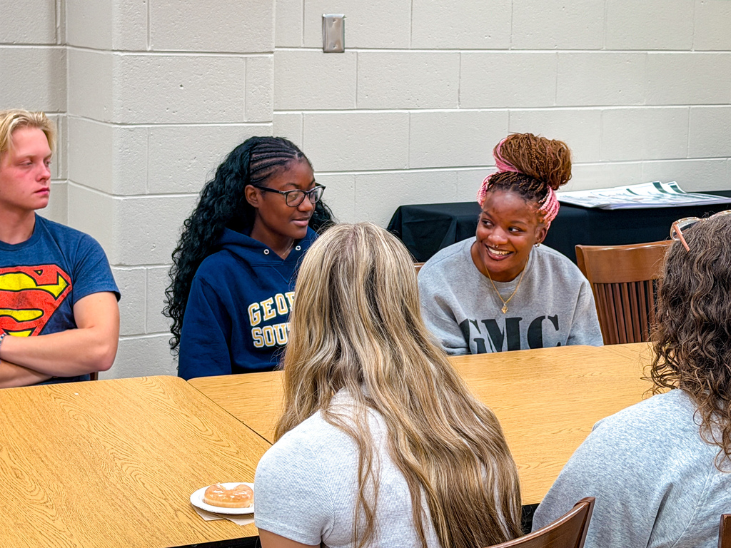 Group of people seated around a table, with one person wearing a Superman t-shirt, and two wearing blue hoodies.