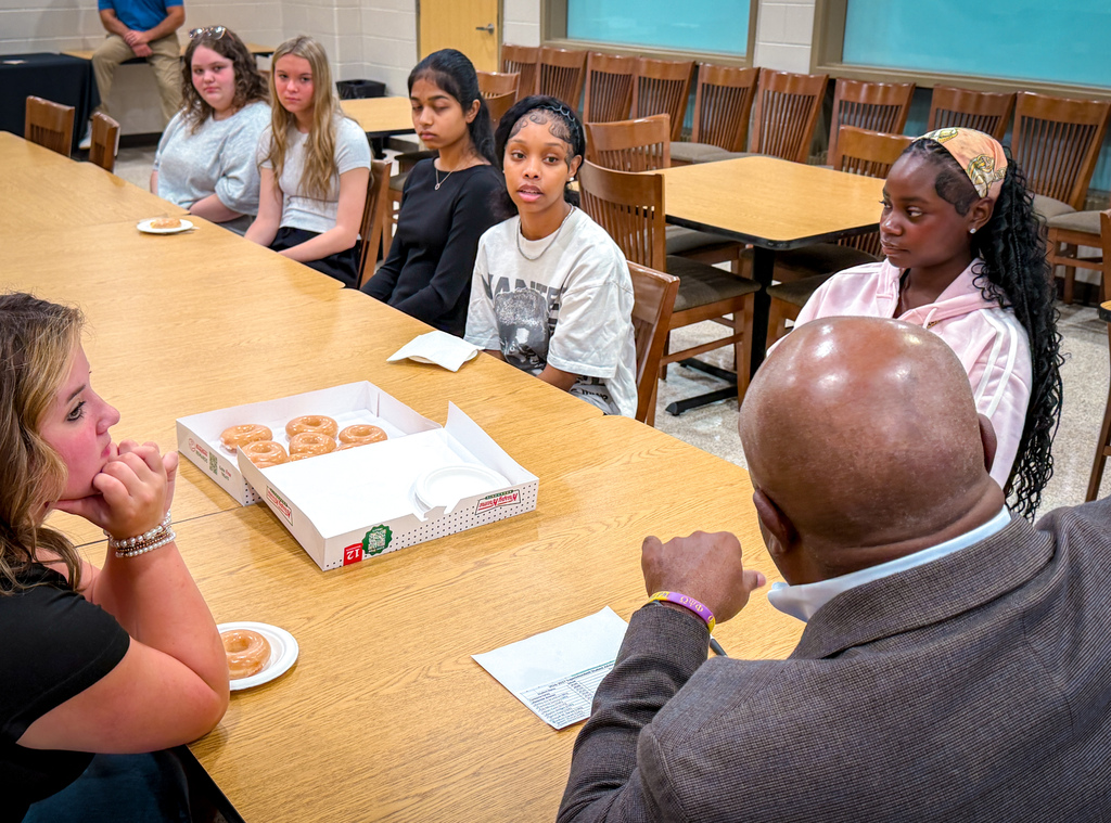A group of individuals seated around a long table, with two donuts on a plate and a box nearby.