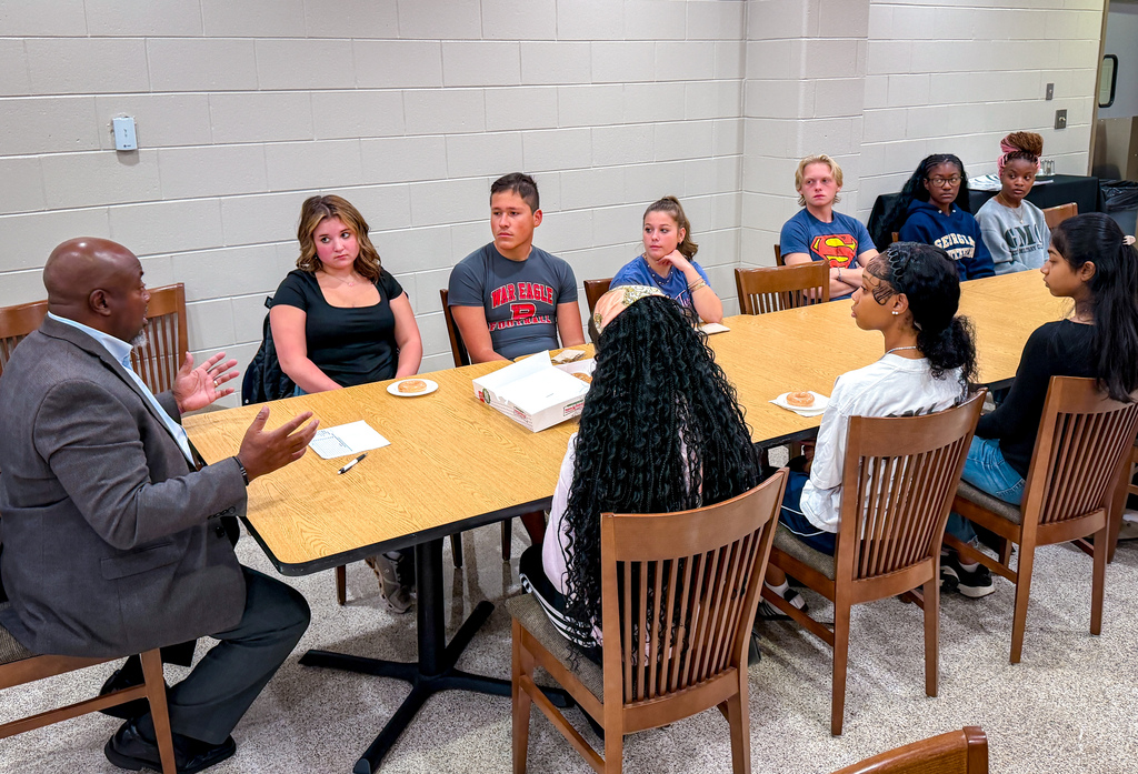 A group of individuals sit around a table in a classroom; a man in a suit speaks while others listen.