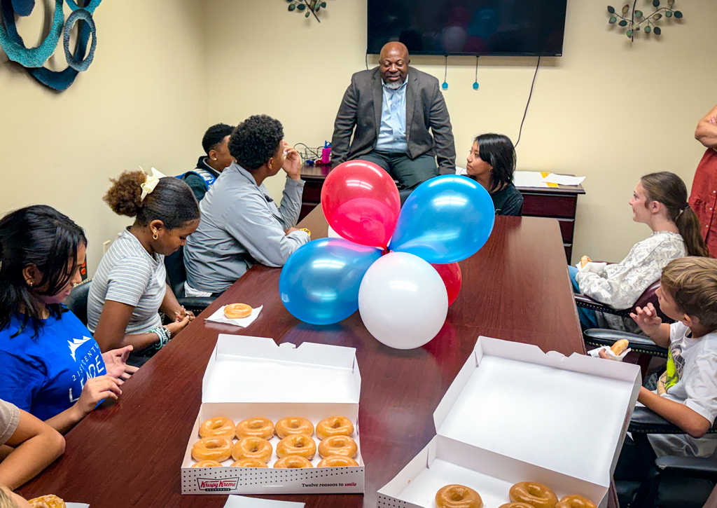 A group of people sit around a table with donuts and balloons. One man stands and speaks to the seated group.