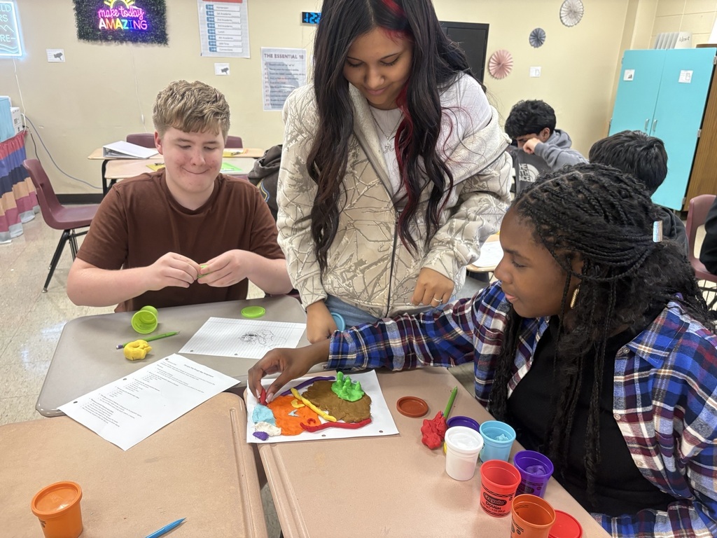 Students are seated at tables with crafts. One student is demonstrating a craft, and two others are observing.