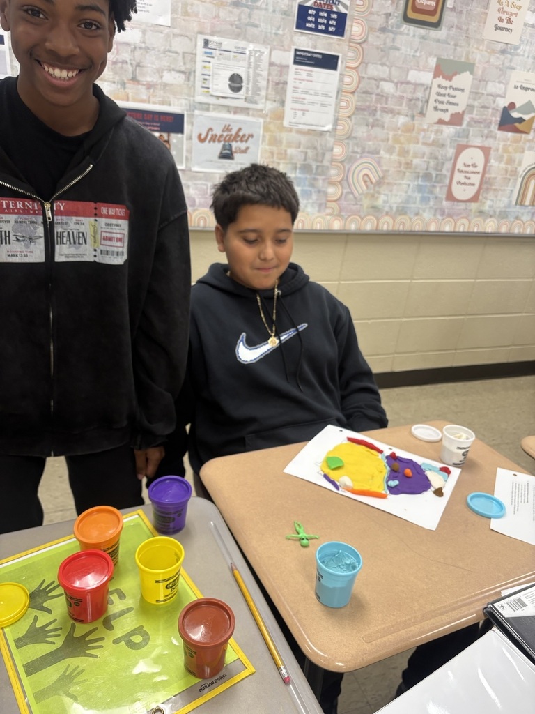 Two boys, one smiling, work on an art project at a table with paint cups and a paper.