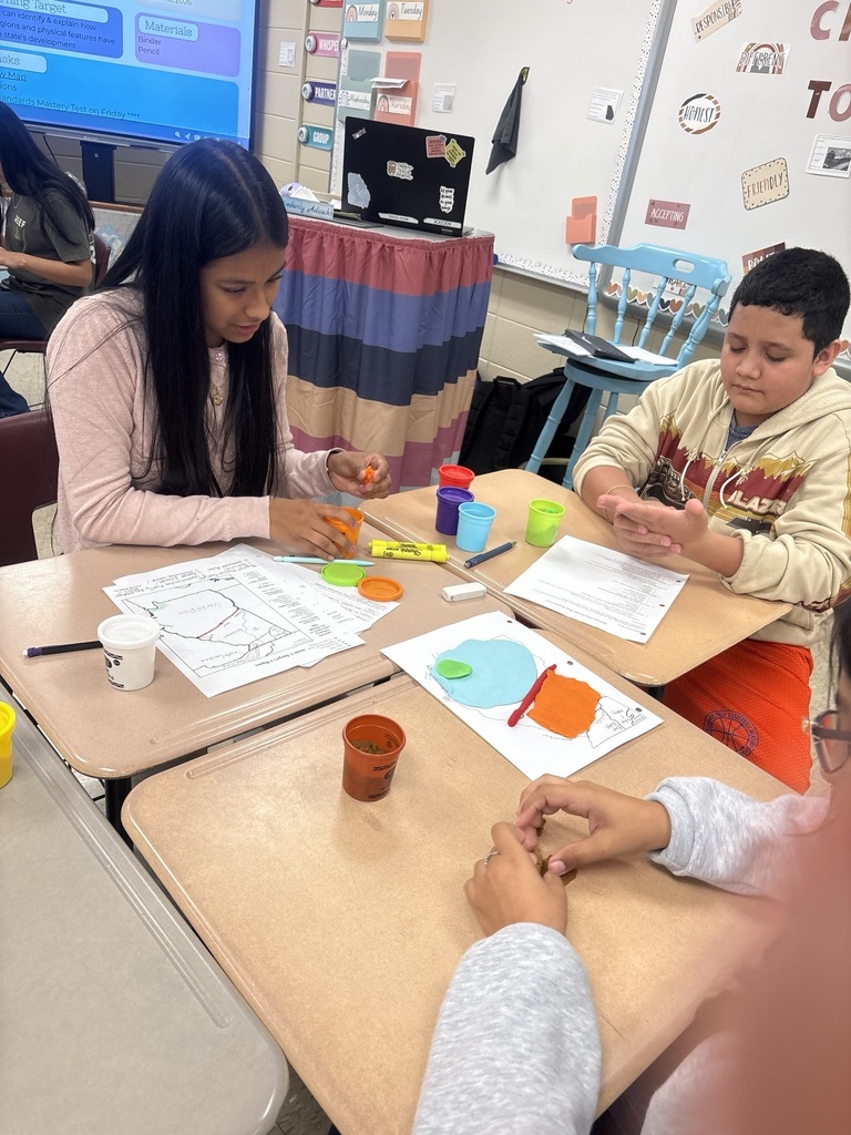 Children seated at desks in a classroom, engaged in a crafting activity using markers and paint, with papers and cups in front of them.