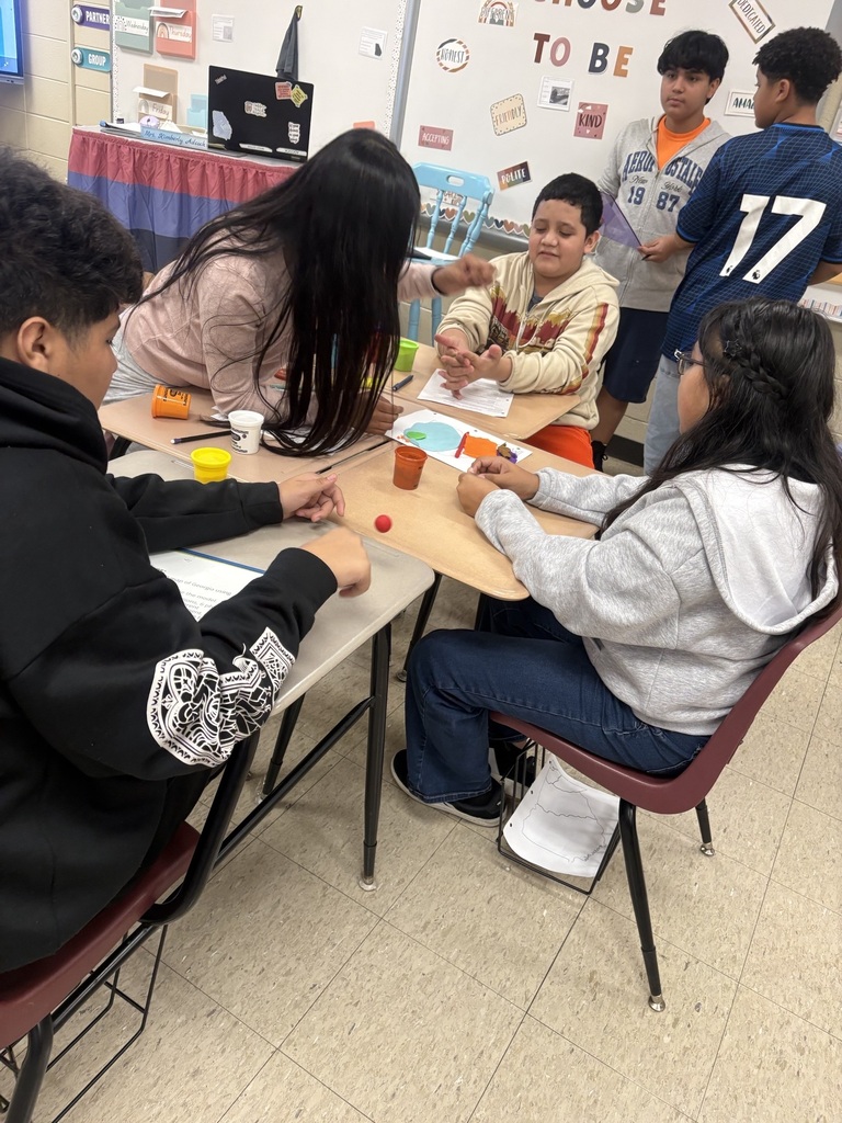 A group of students work on a project at tables in a classroom, with a whiteboard in the background.