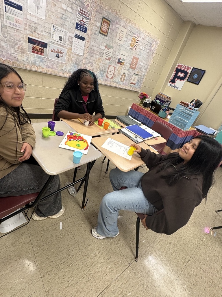 Three people in a classroom with a bulletin board. One sits at a table with a colorful pie chart.