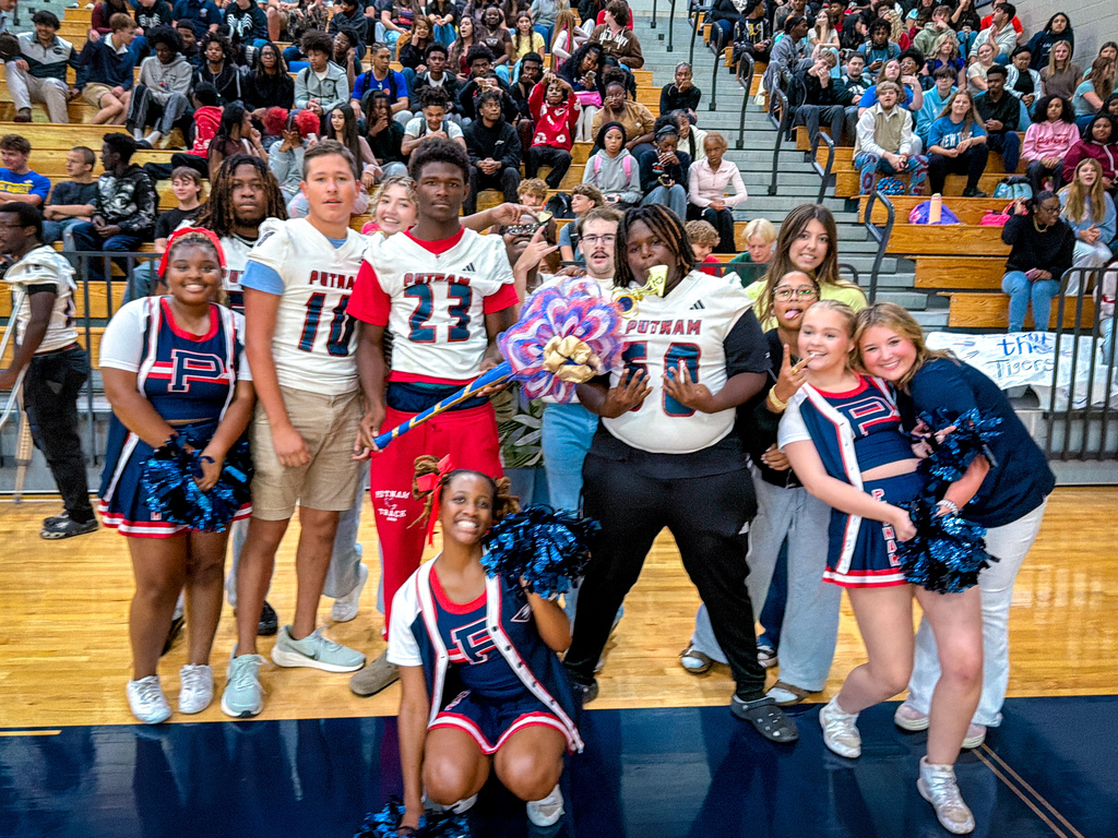 Group of athletes and cheerleaders in blue uniforms standing together on a basketball court, with spectators in the background.