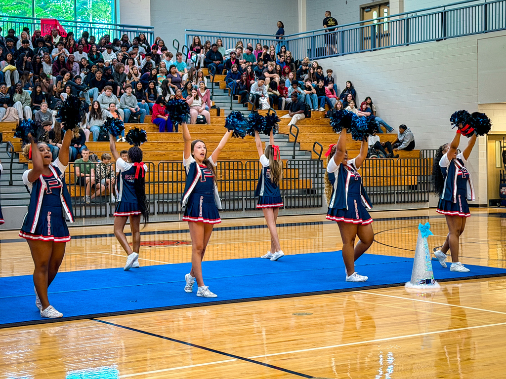 A group of cheerleaders performing on a blue mat in a gym. The audience is seated in the background.