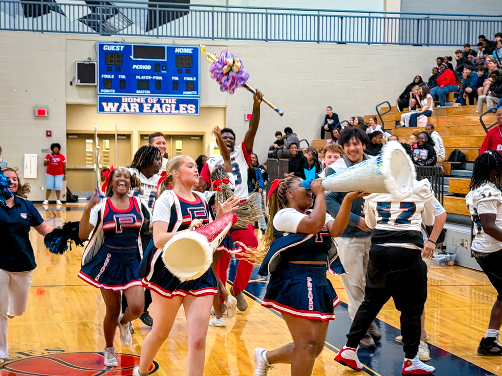 Cheerleaders run across a court in front of an audience. One holds a pompom and megaphone.