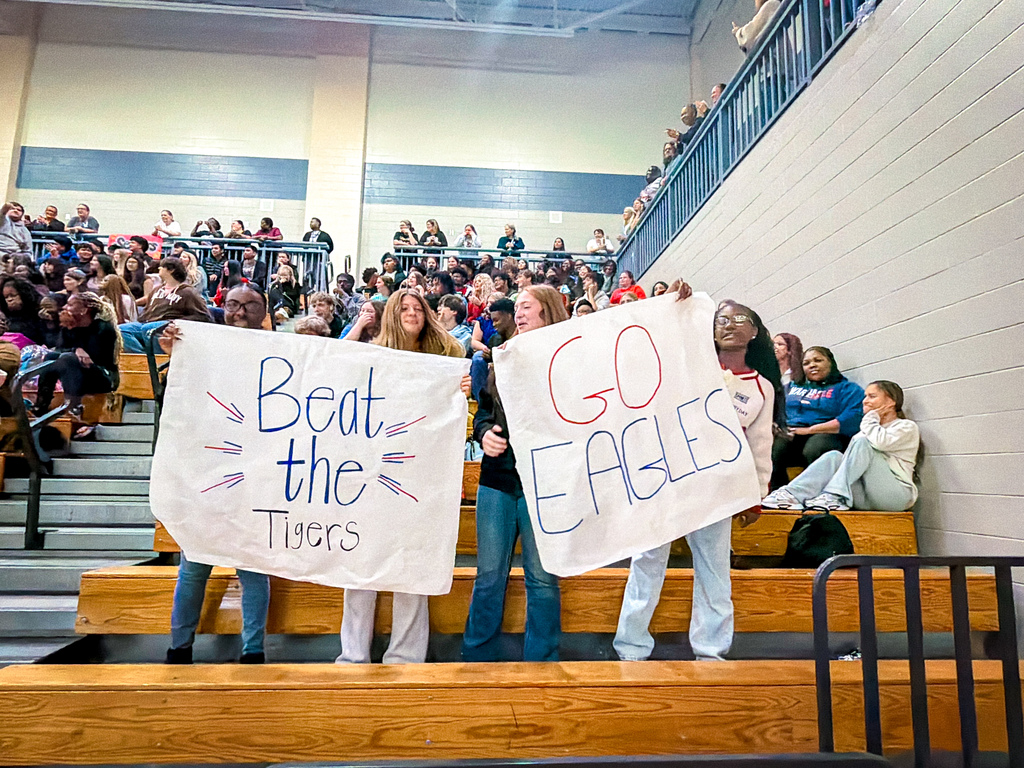 People in a sports arena holding signs with "Beat the Tigers" and "Go Eagles" messages, seated in bleachers.