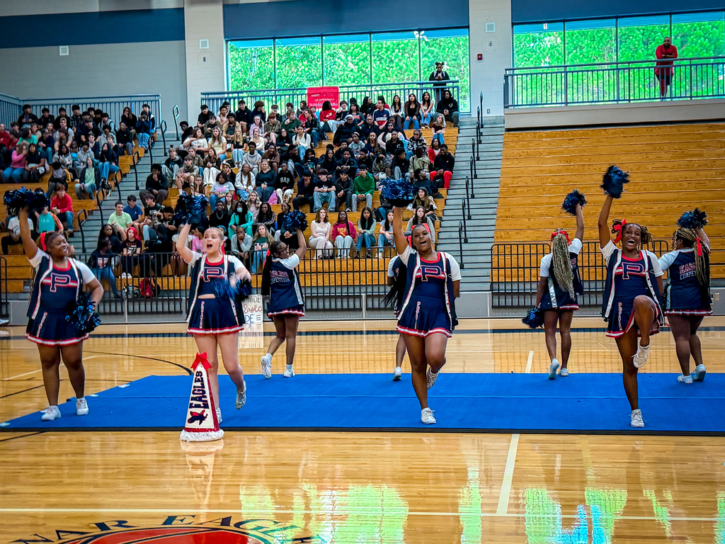 Cheerleaders in blue uniforms perform on a blue mat on a basketball court, with an audience seated in the stands.