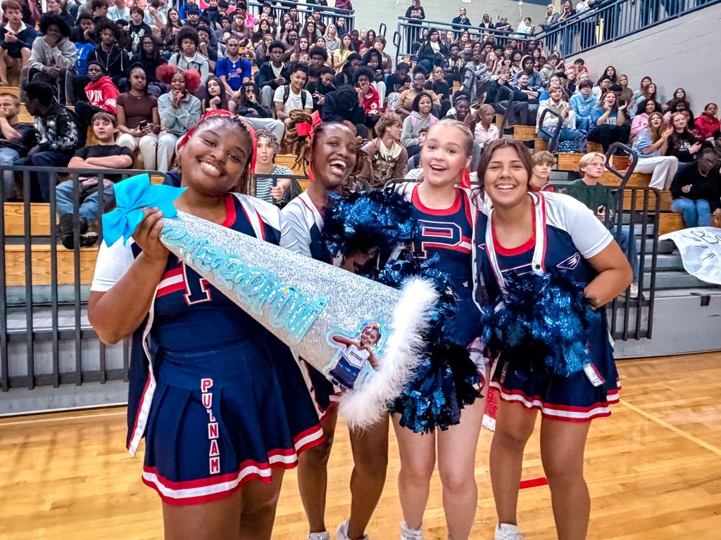 Four cheerleaders in blue uniforms pose on a basketball court with a crowd in the stands.
