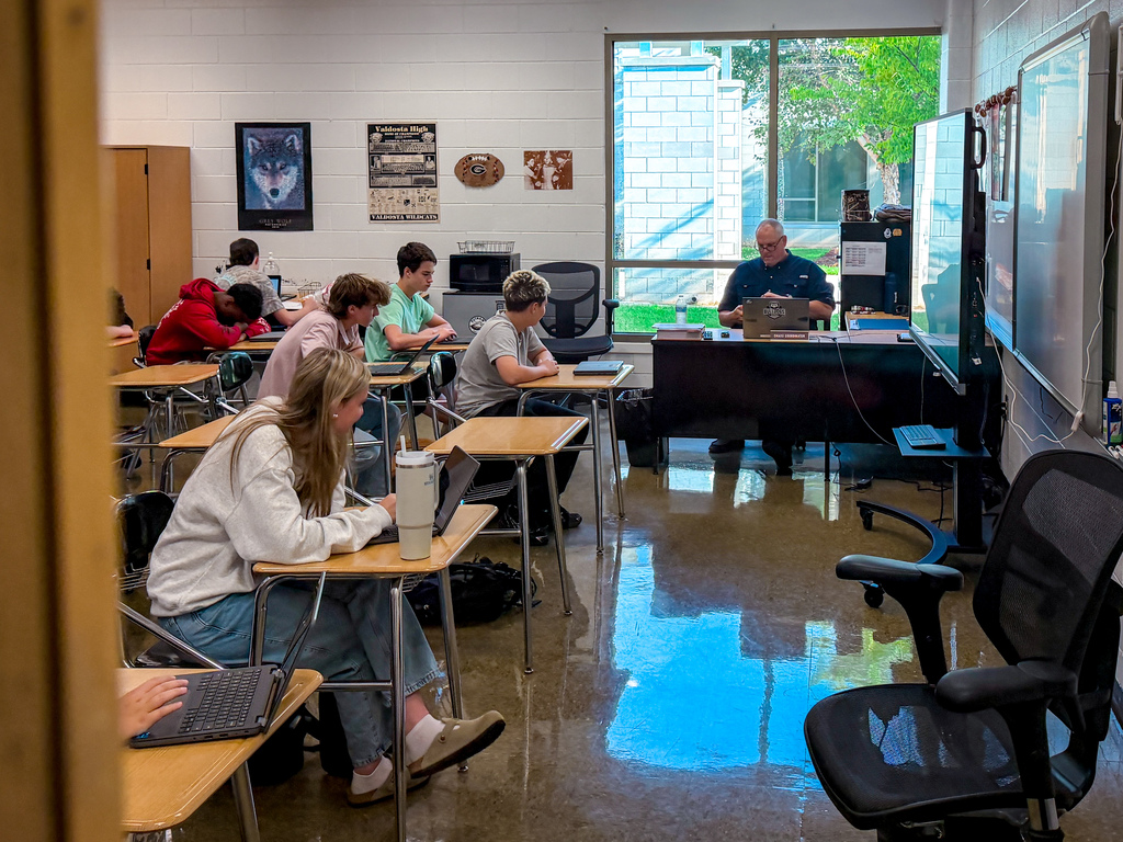 A classroom with students seated at desks, using laptops, a teacher at a desk with a monitor, and two black office chairs.