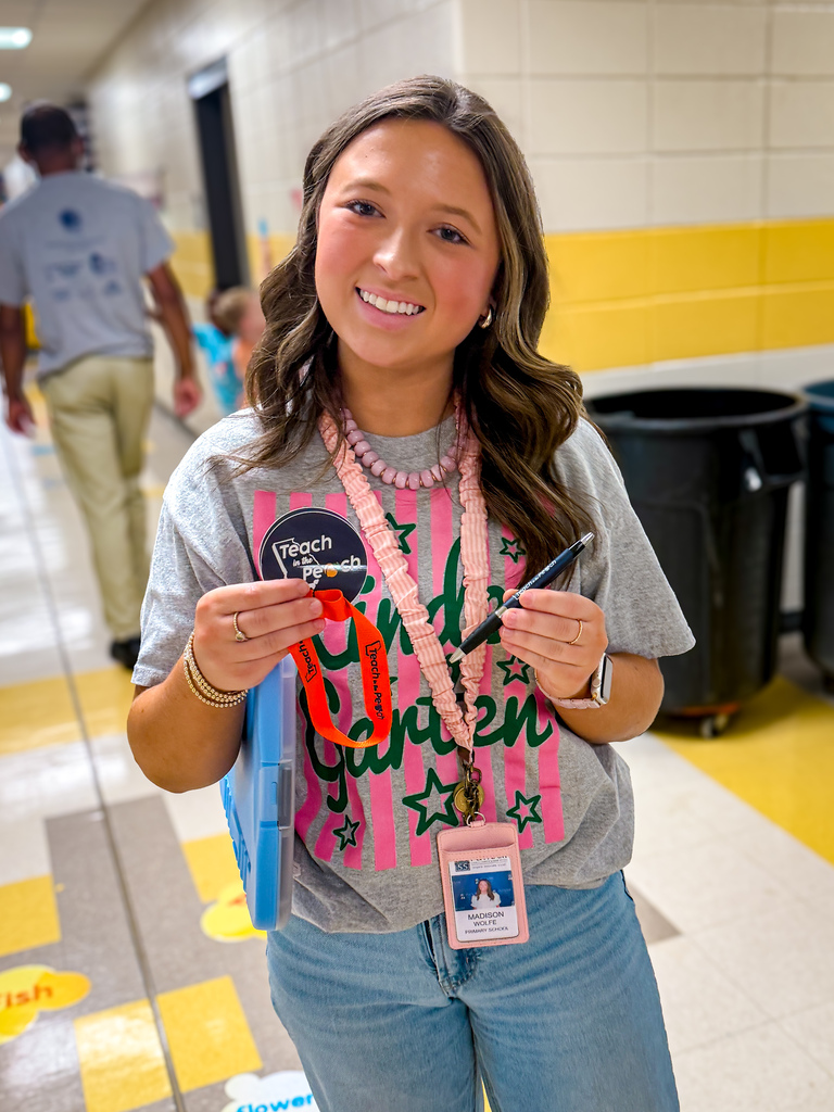 A woman holds a badge and pen, smiling in a school hallway with yellow and white tiles.