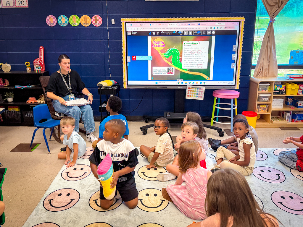 Kids sit on a rug with smiling faces, watching a teacher on a screen in a classroom.