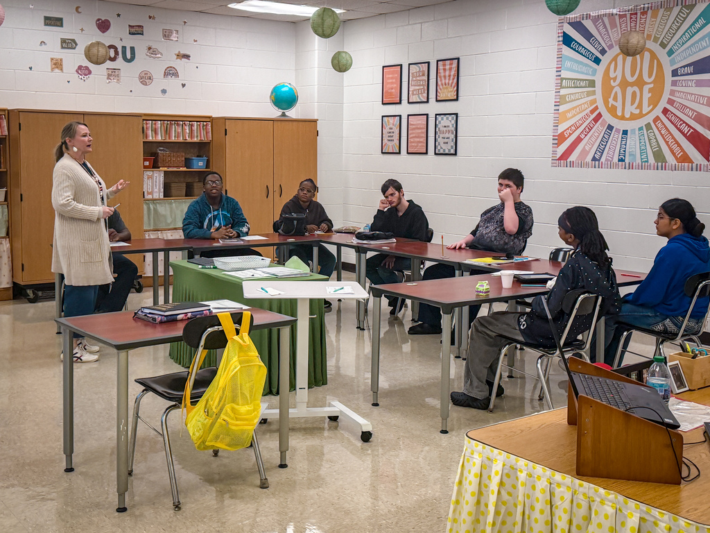 A classroom with students sitting at tables and a teacher standing in front of them.