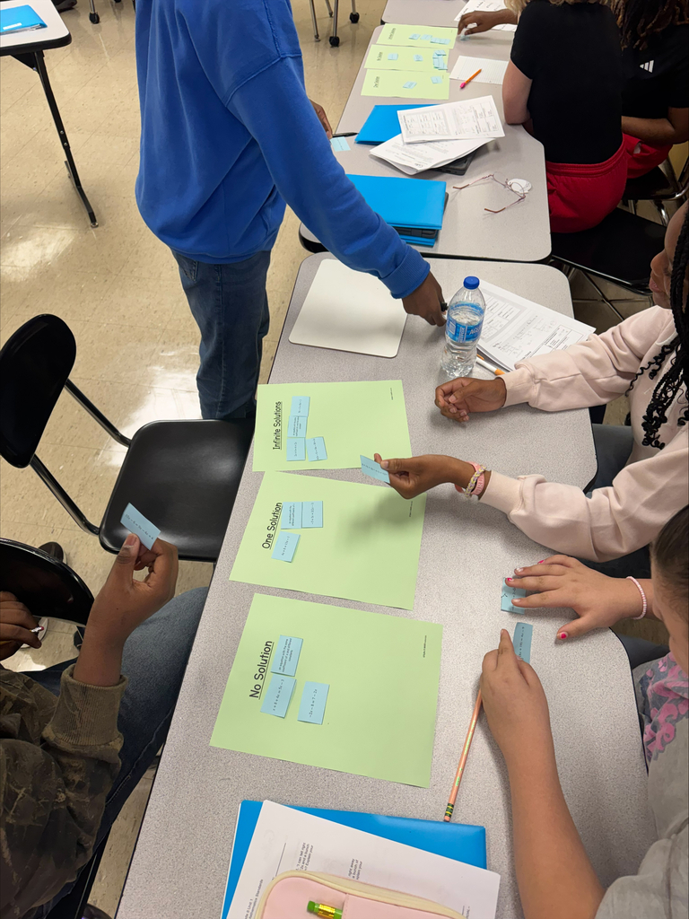 Students seated at a long table, engaging in a classroom activity with colored papers, markers, and other tools.