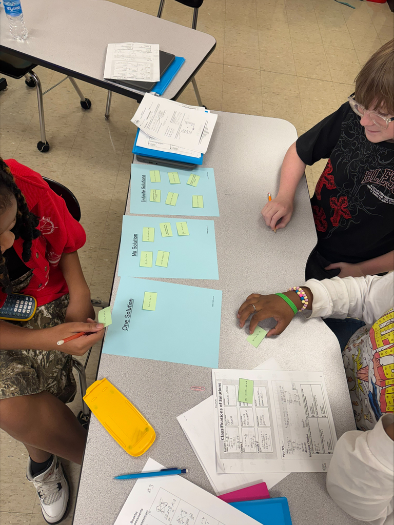 Three students at a table, each holding a yellow sticky note, engaged in a group activity.