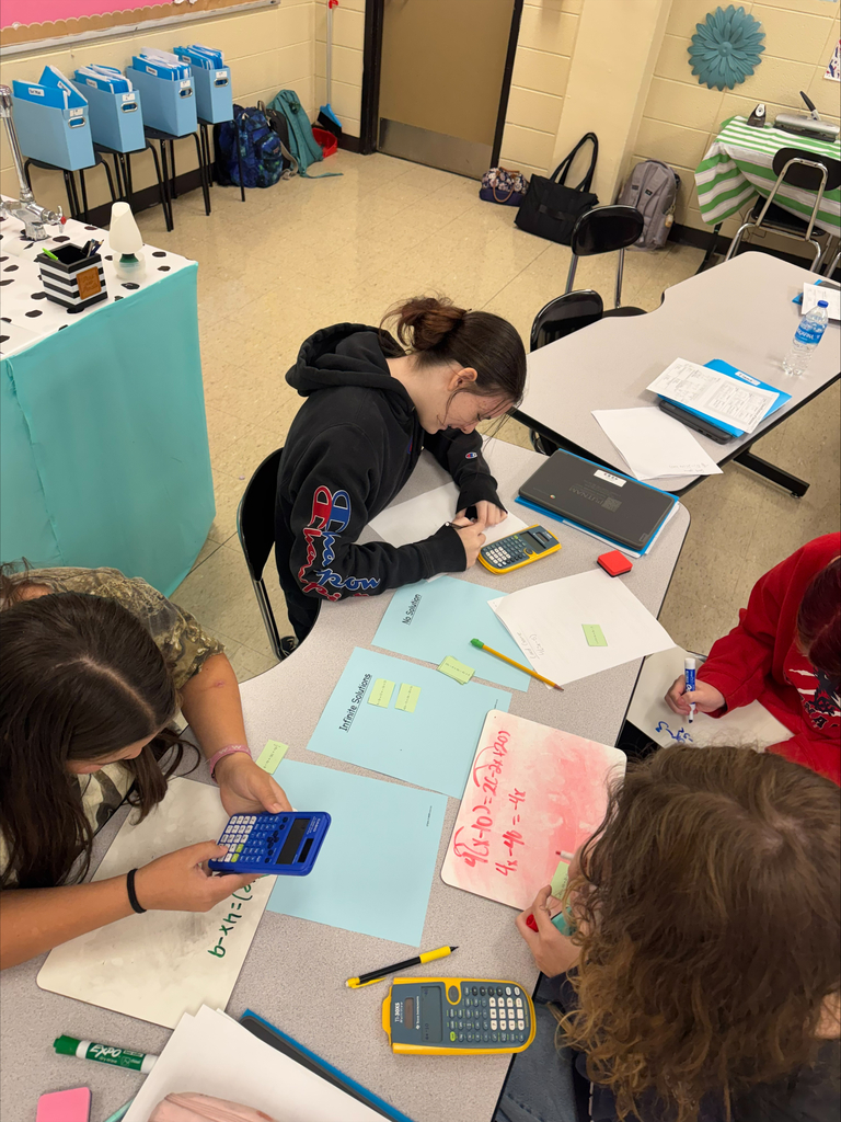 Four students seated at a table, engaged in writing on paper with calculators, pens, and markers around them.
