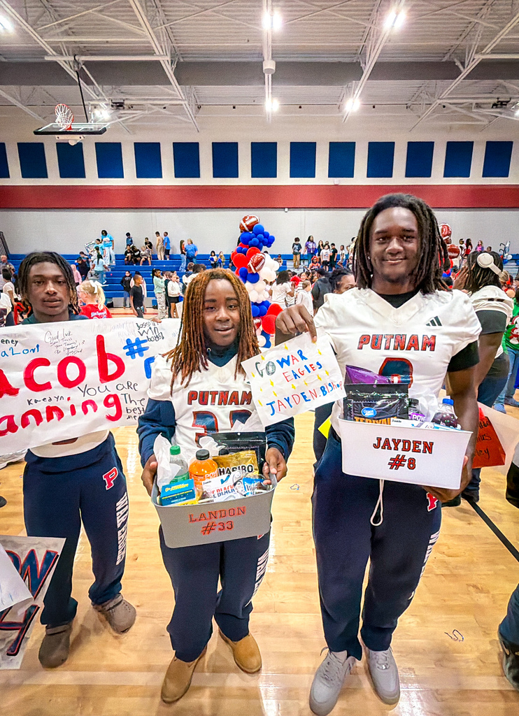 Three students hold boxes with team jerseys and gifts, standing in a gym with a crowd in the background.
