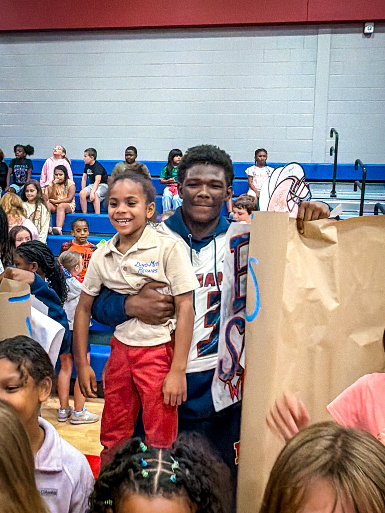 A man and a child in red shorts hold a brown paper bag in a gym. Several children watch from the bleachers.