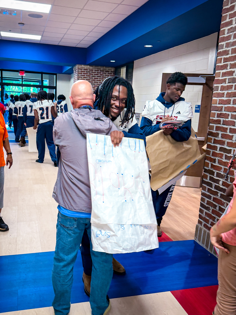 A man in gray jacket and jeans holds a white paper while hugging another man in blue.
