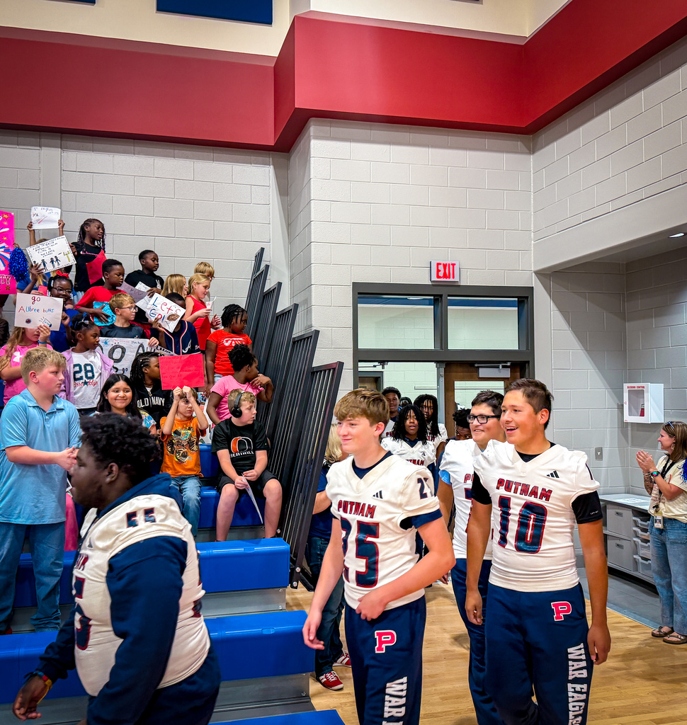 Three young men in football uniforms walk past a crowd in a gymnasium. Some people hold up signs.