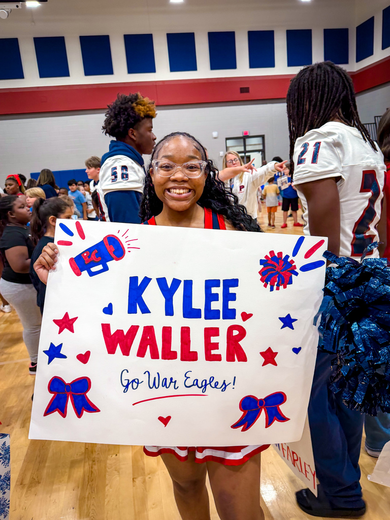 Cheerleader holds a sign for Kylee Waller and War Eagles while other people in sports jerseys walk around.