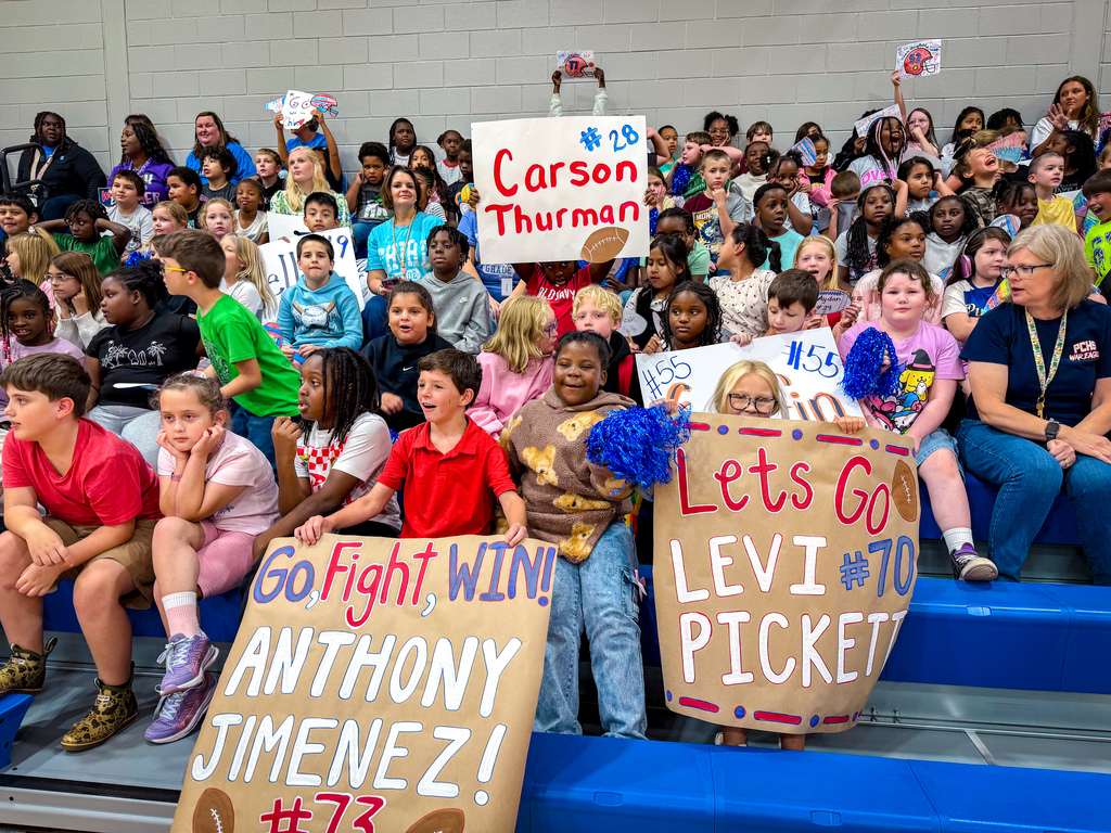 Many children in a gym, some standing, holding signs that read "Carson Thurman," "Anthony Jimenez," and "Levi Pickett."