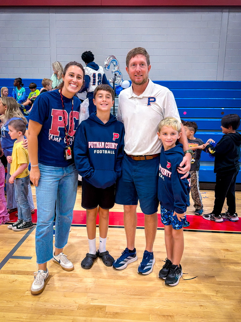 A family of four, wearing matching blue hoodies, stands in a gymnasium with a mascot. Children in the background watch.