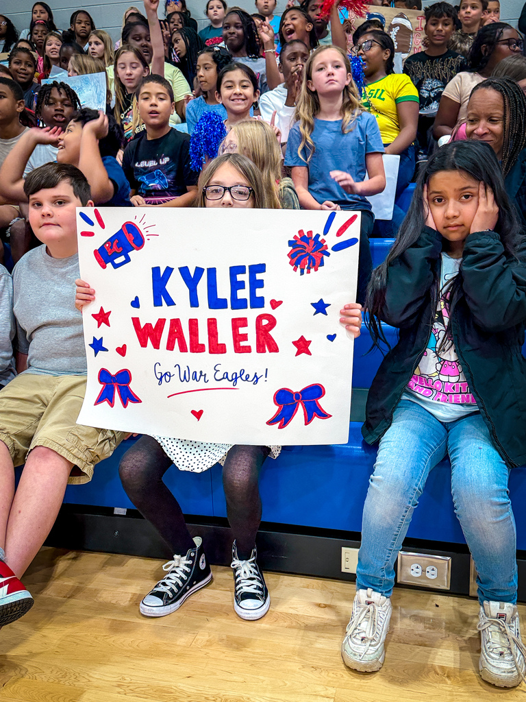 A group of children seated on a blue bench. One child holds a sign that reads "Kylee Waller Go War Eagles!"