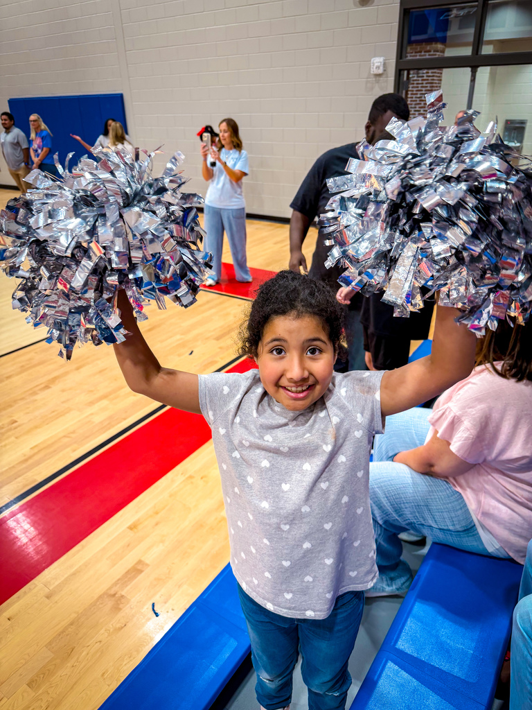 A young girl holds silver pompoms above her head on a blue bench in a gym.