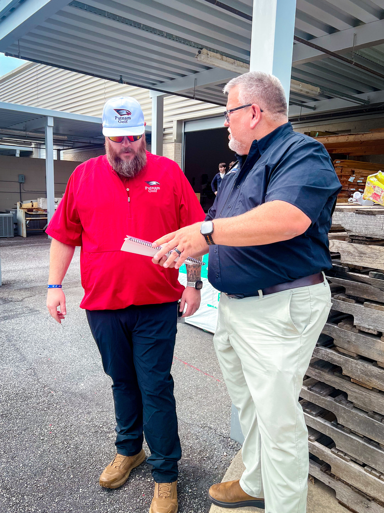 Two men, one in red and one in blue, stand outdoors. One holds a paper while the other points. A wooden pallet stack is behind them.