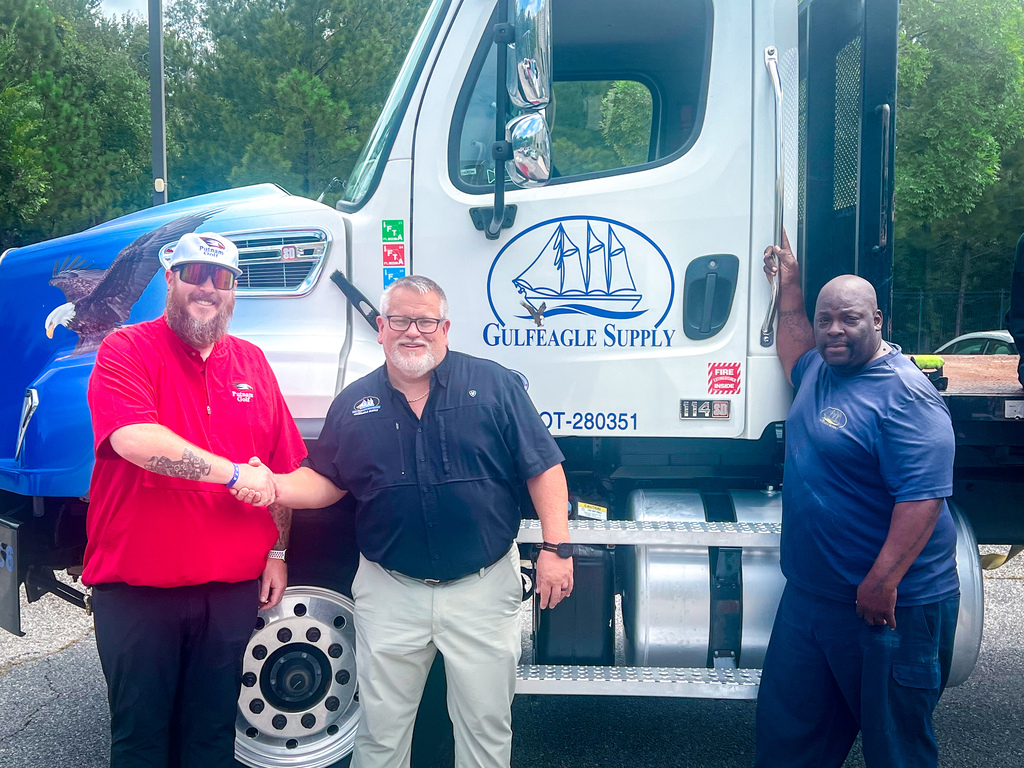 Three men in casual clothes stand before a large white truck with a blue logo, shaking hands.