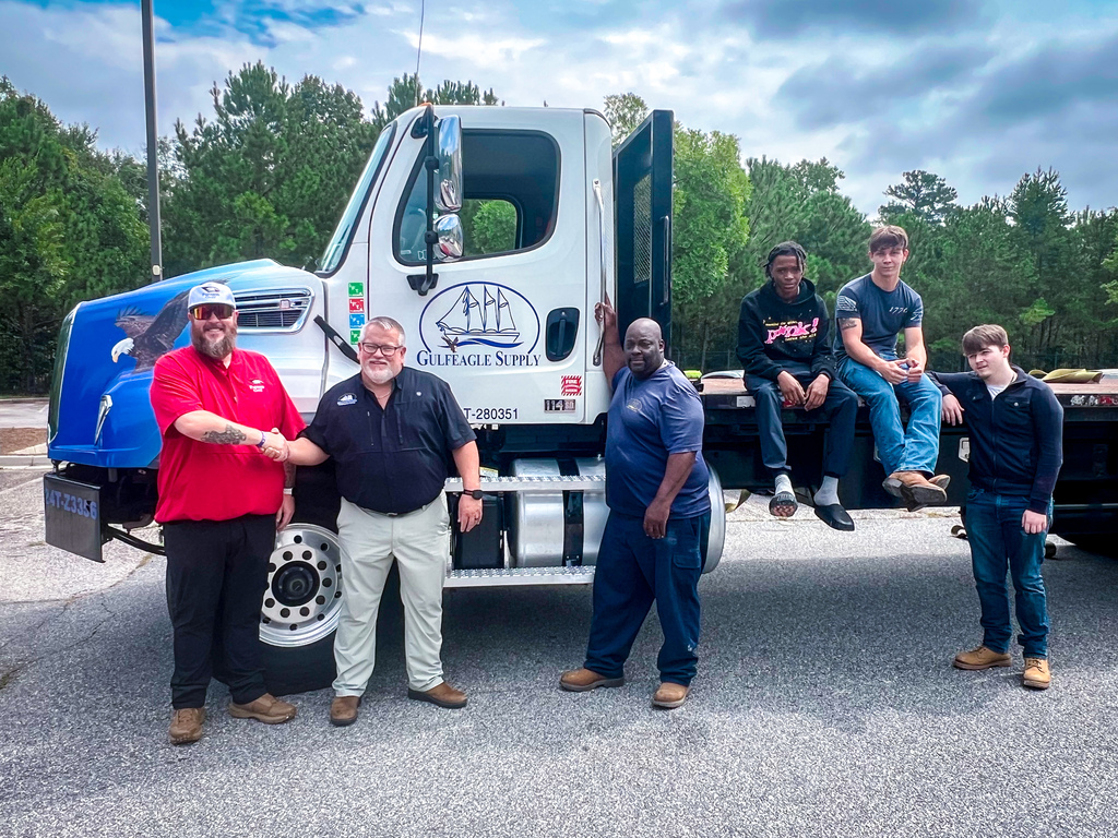 Five men stand in front of a large white truck with a blue design. Three sit on the truck bed, while two stand in front.