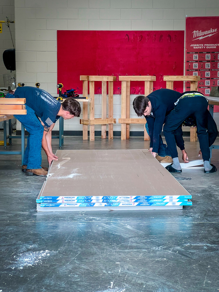 Two people are working on a project in a workshop with wooden stools and a red wall in the background.