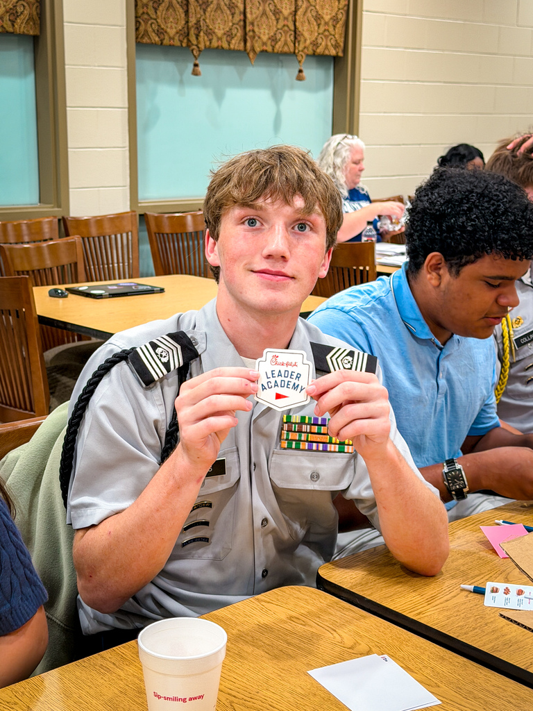 Person in gray military-style uniform holds card with "Leader Academy" text. Seated at table with others.